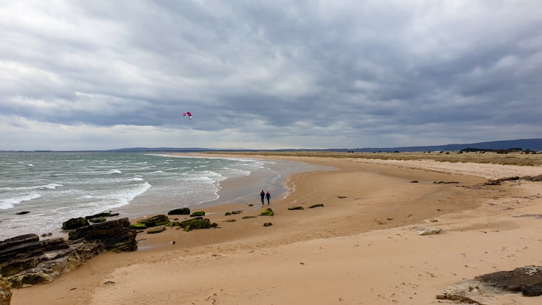 A couple walking along Dornoch Beach with a kite surfer in the distance